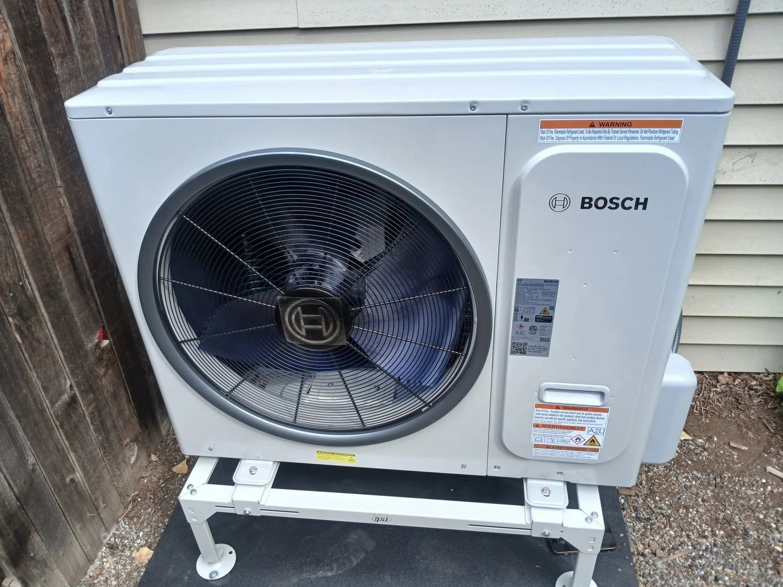 A Bosch heat pump on a white stand beside a wood fence