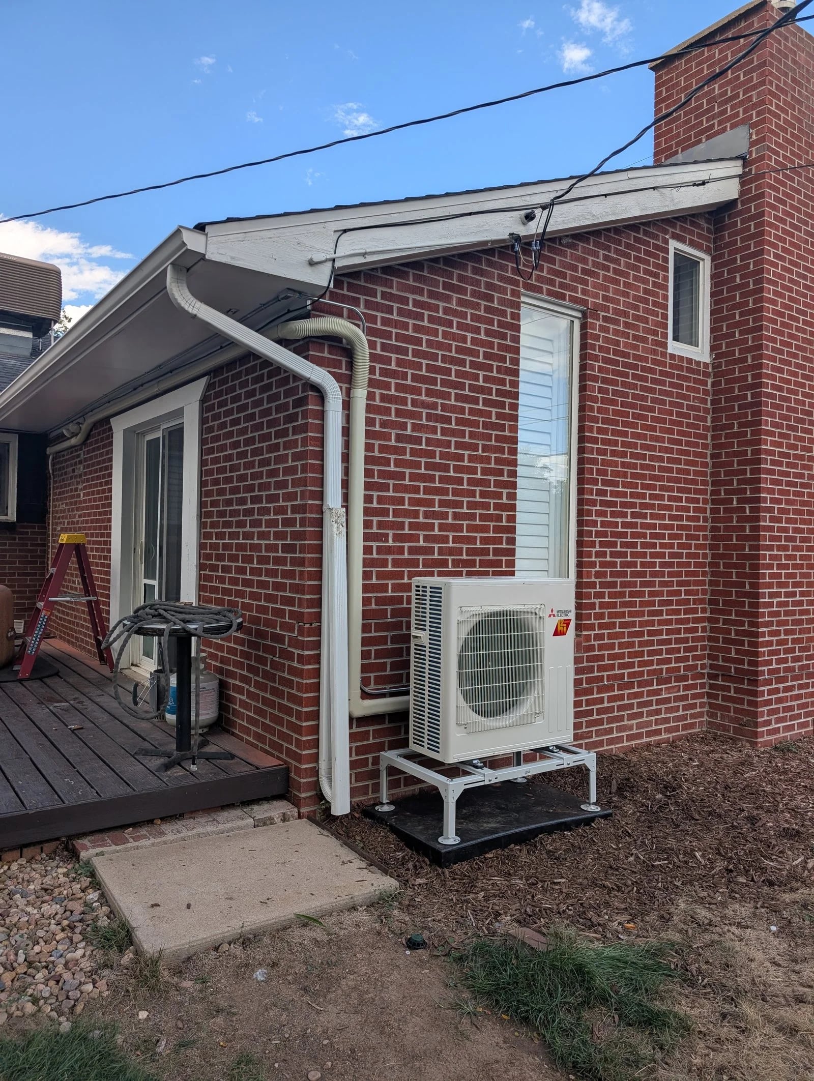 A heat pump on a stand beside a red brick bungalow with a small wood deck