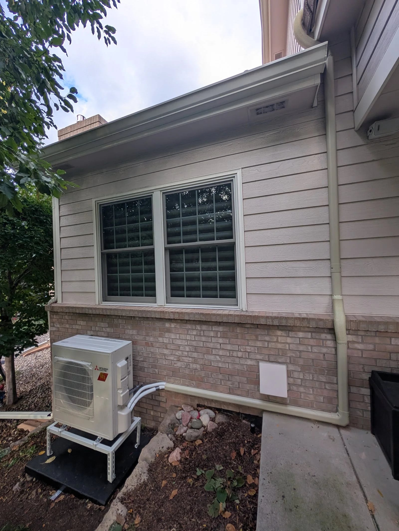 A heat pump on a stand below a window, with a line-set cover running up the siding