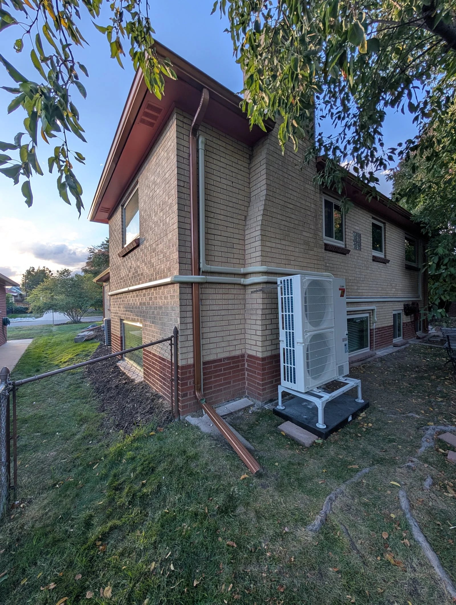 A multi-zone heat pump on a stand beside a brick two-story house, with line-set covers up the wall
