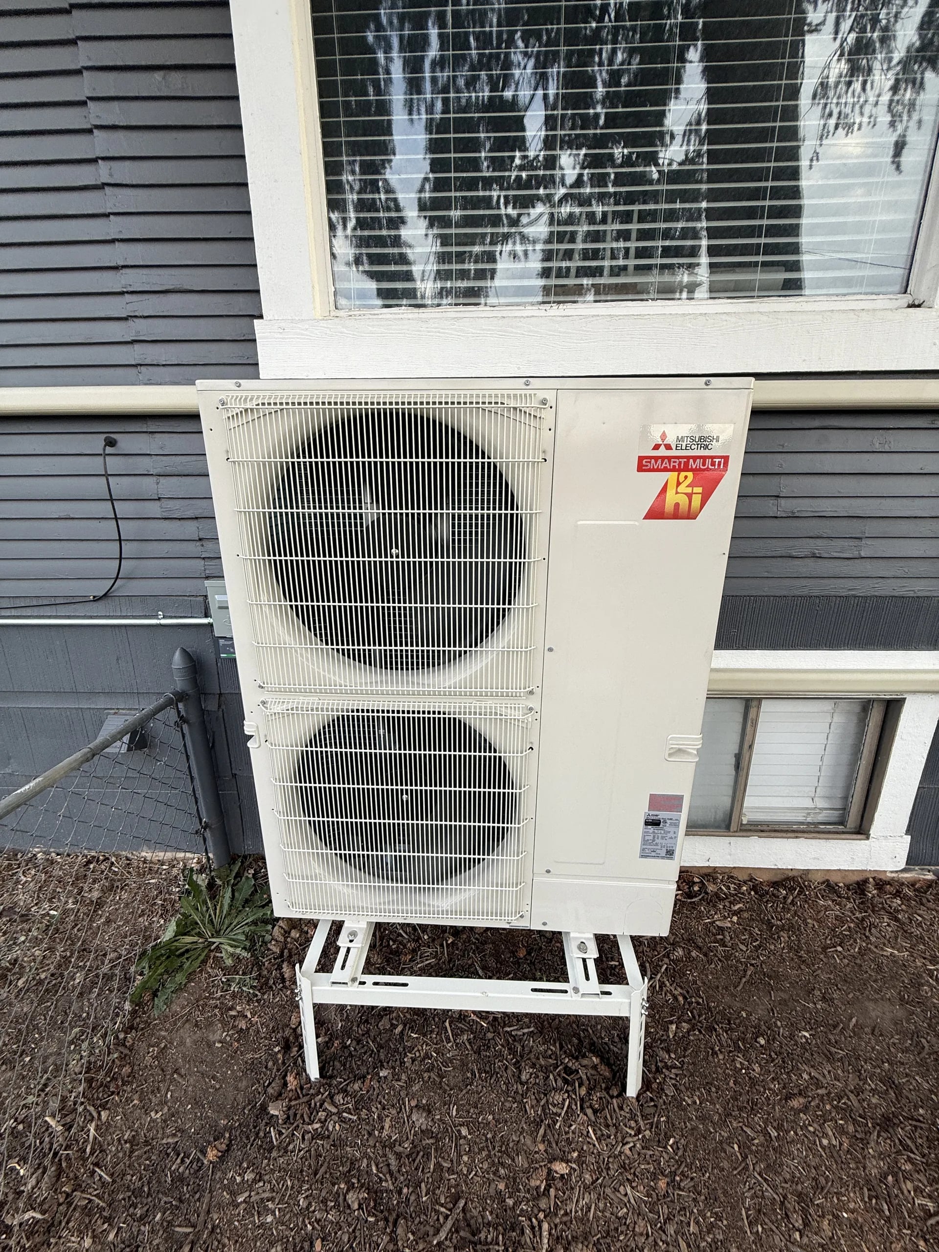 Mitsubishi two-fan outdoor unit on a white stand in mulch below a window on gray siding